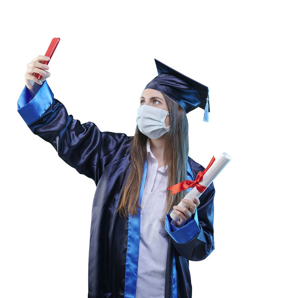 Menina graduada está tirando uma foto de si mesmo, pandemia e estilo máscara com boné e vestido, mostrando certificado. Fundo colorido. - Foto, Imagem