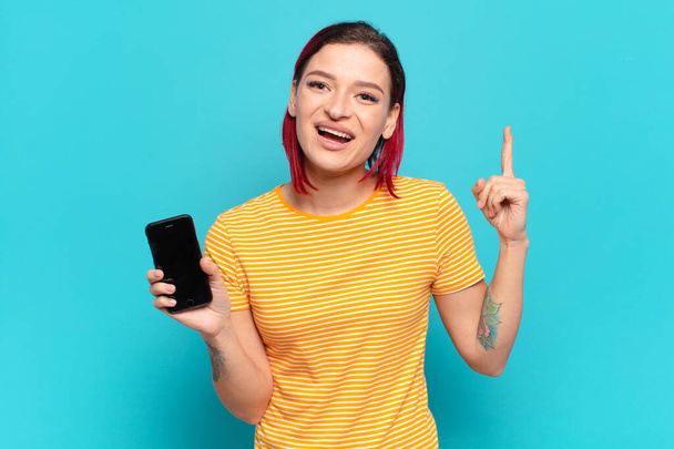 young attractive red hair woman feeling like a happy and excited genius after realizing an idea, cheerfully raising finger, eureka! and showing her cell - Foto, imagen