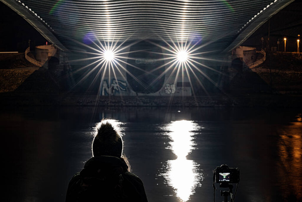 Silhouette di una donna con un berretto in testa e una macchina fotografica su un treppiede che guarda il livello del fiume di notte. - Foto, immagini