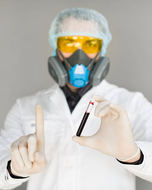 Függőleges fotó egy kutatóról orvosi köpenyben és védőmaszkban, kezében egy kémcsővel, szennyezett vérrel a kezében. A modern kutatás koncepciója a vakcinafejlesztés területén. - Fotó, kép