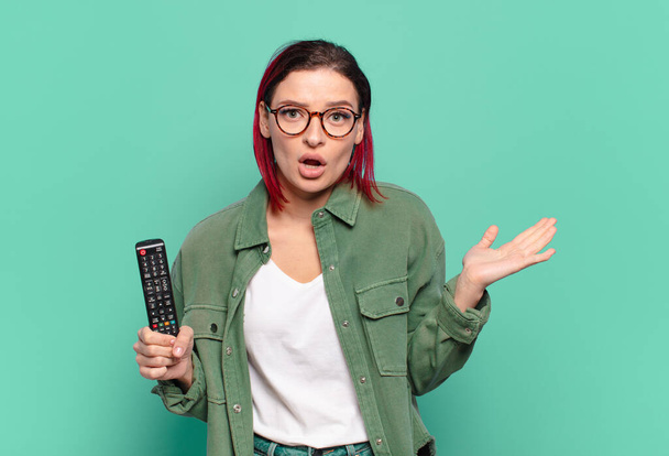 young attractive red hair woman looking surprised and shocked, with jaw dropped holding an object with an open hand on the side and holding a tv remote control - Photo, Image