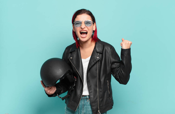 young attractive red hair woman shouting aggressively with an angry expression or with fists clenched celebrating success. motorbike rider concept - Photo, Image