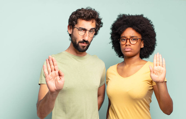 multiracial couple of friends looking serious, stern, displeased and angry showing open palm making stop gesture - Fotoğraf, Görsel