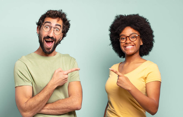 multiracial couple of friends looking excited and surprised pointing to the side and upwards to copy space - Фото, изображение