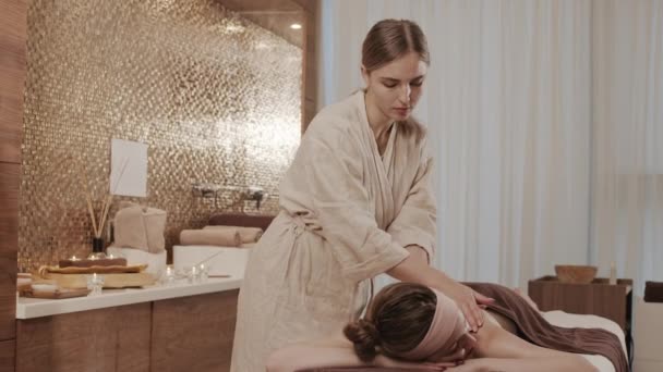 Colpo medio di giovane massaggiatrice che indossa accappatoio di lino facendo ringiovanire massaggio alla schiena per giovane donna attraente sdraiata sul tavolo da massaggio nel centro benessere - Filmati, video
