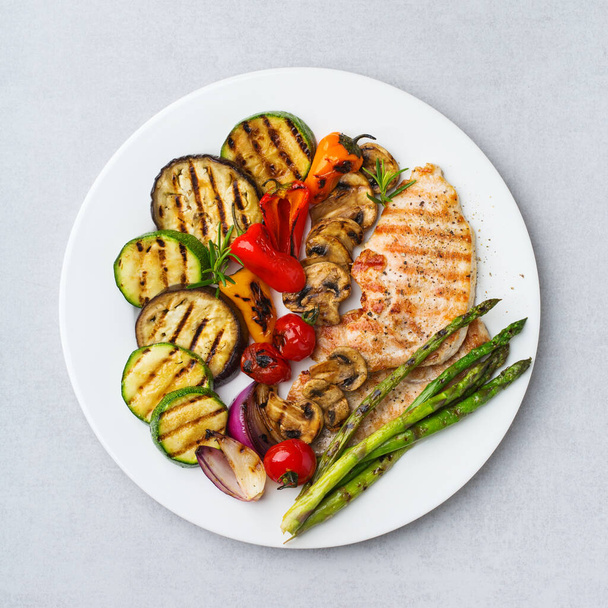 Saisonales, sommerliches Essenskonzept. Gegrilltes Gemüse und Hühnerbrust in einem Teller auf einem weißen Tisch. Draufsicht flacher Hintergrund - Foto, Bild