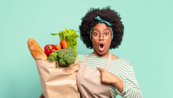 joven bastante afro mujer buscando sorprendido y sorprendido con la boca abierta, apuntando a sí mismo y sosteniendo una bolsa de verduras - Foto, Imagen