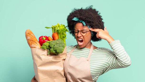 young pretty afro woman looking unhappy and stressed, suicide gesture making gun sign with hand, pointing to head and holding a vegetables bag - 写真・画像