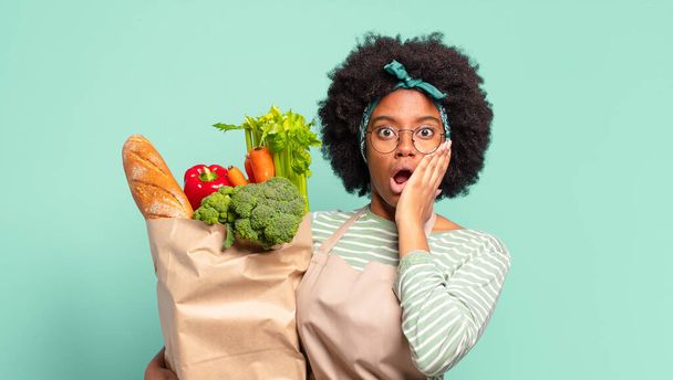 young pretty afro woman feeling shocked and scared, looking terrified with open mouth and hands on cheeks and holding a vegetables bag - Foto, Bild