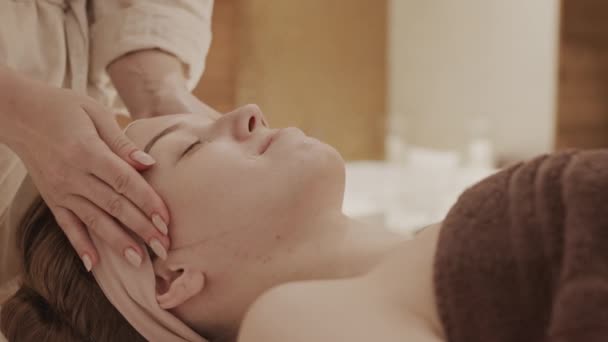Зв'язок нерозпізнаної жінки-косметолога з використанням зволожувального крему та масажу для обличчя молодої кавказької жінки, що лежить на масажному столі в спа-салоні. - Кадри, відео