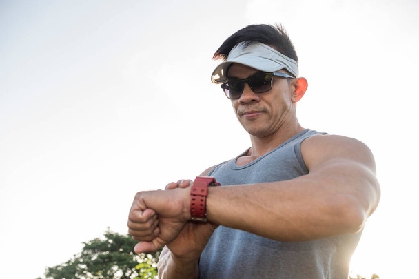 A fit asian man checks the time, or data like pulse, heart rate, distance or calories burned on his smartwatch. Outdoor scene. - Photo, Image