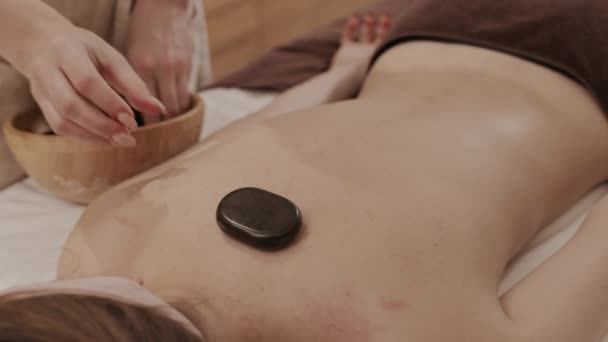 Vue grand angle de femme esthéticienne méconnaissable mettant des pierres noires chaudes sur le dos d'une jeune femme méconnaissable allongée sur une table de massage et relaxante - Séquence, vidéo