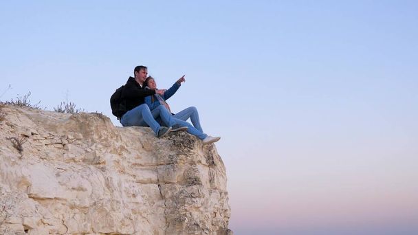 Bezpłatny podróżnik kobieta i mężczyzna siedzą na krawędzi górskiego przytulania i ciesząc się pięknym krajobrazem. Rodzina turystów spoczywa na skraju klifu. Turyści na szczycie klifu przeciwko błękitnemu niebu - Zdjęcie, obraz