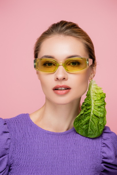 mujer joven en gafas de color y pendientes de lechuga fresca aislada en rosa - Foto, Imagen
