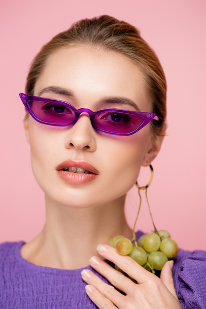 charming woman in purple eyeglasses, with fresh grapes earring isolated on pink - Photo, Image