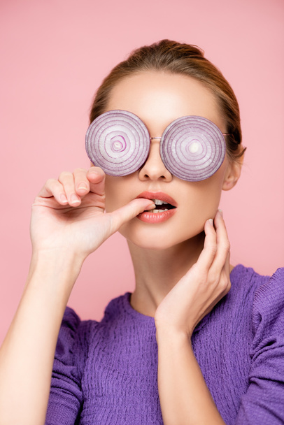 чувственная женщина в очках с фиолетовыми луковыми кольцами, кусающий палец изолирован на розовый, сюрреализм концепции - Фото, изображение