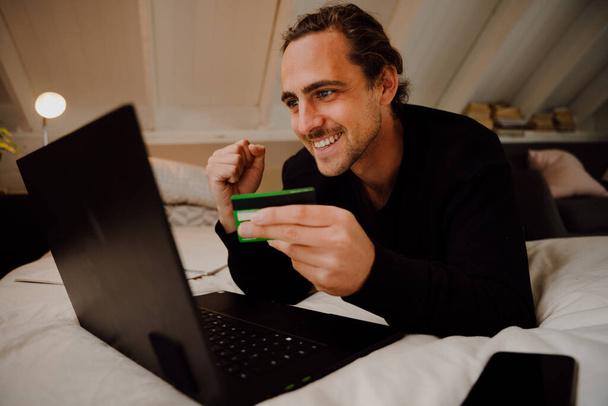 Feliz caucásico estudiante masculino con éxito haciendo el pago en línea utilizando la escritura de tarjetas de crédito en el ordenador portátil - Foto, Imagen