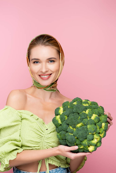 happy woman in kerchief holding green broccoli while smiling at camera isolated on pink, surrealism concept - Foto, Imagen