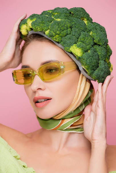 giovane donna in occhiali colorati cappello toccante fatto di broccoli freschi isolati su rosa, concetto di surrealismo - Foto, immagini