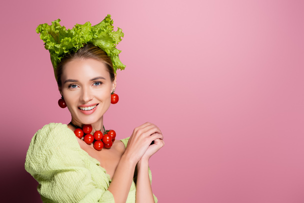 smiling woman in hat, necklace and earrings made of vegetables posing on pink - Foto, afbeelding