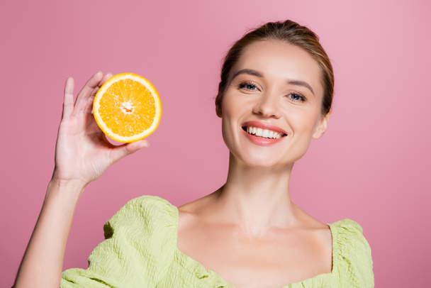 joyful woman with natural makeup holding half of fresh orange isolated on pink - 写真・画像