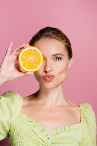 pretty woman sending air kiss while covering eye with orange half on pink - Photo, Image