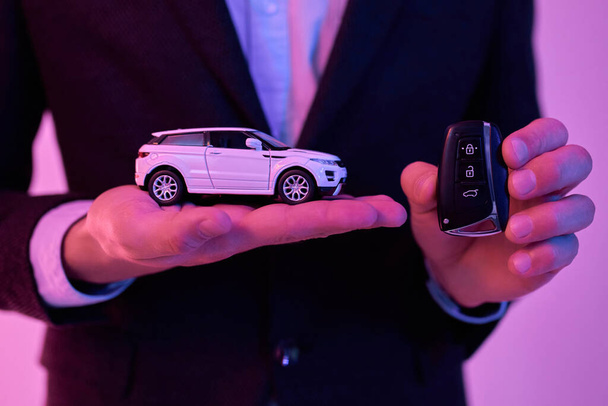 Foto ritagliata di un giovane uomo d'affari attraente in un abito nero e camicia bianca che tiene un modello di auto nel palmo della mano, isolato su uno sfondo rosa fluo. Acquisto auto, noleggio. - Foto, immagini