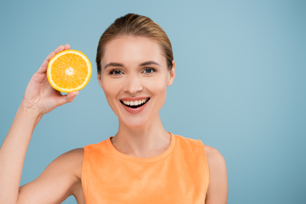 radostná žena se usmívá na kameru, zatímco drží polovinu šťavnaté pomeranče izolované na modré - Fotografie, Obrázek