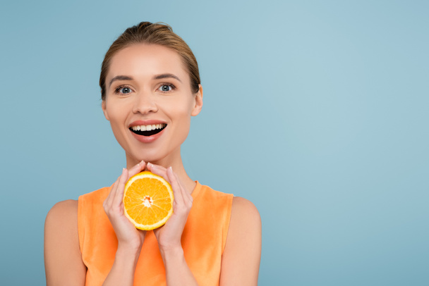 mujer sorprendida con maquillaje natural sosteniendo la mitad de naranja jugosa aislada en azul - Foto, Imagen