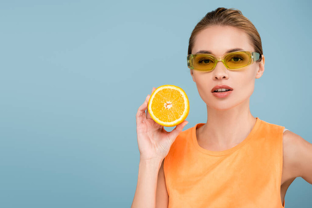 charming woman in colored eyeglasses posing with half of ripe orange isolated on blue - Photo, Image