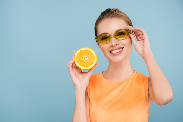 cheerful woman touching colorful eyeglasses while holding juicy orange isolated on blue - Foto, Imagen