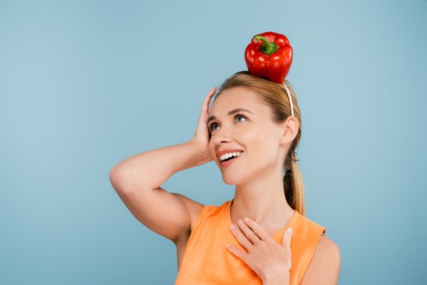 excited woman with red bell pepper instead of hat posing with hand on chest isolated on blue - Photo, Image