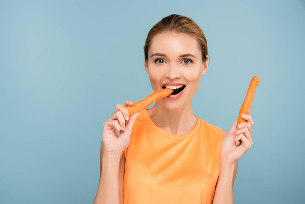 pleased woman looking at camera while eating whole carrot isolated on blue - Photo, Image