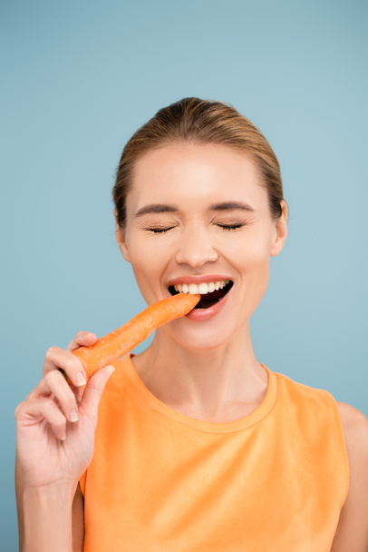 alegre joven con maquillaje natural mordiendo zanahoria entera aislada en azul - Foto, imagen