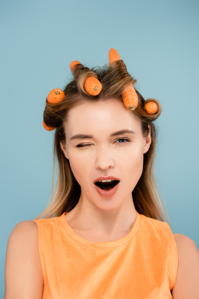 flirty woman with carrots instead of curlers winking at camera isolated on blue - Foto, imagen