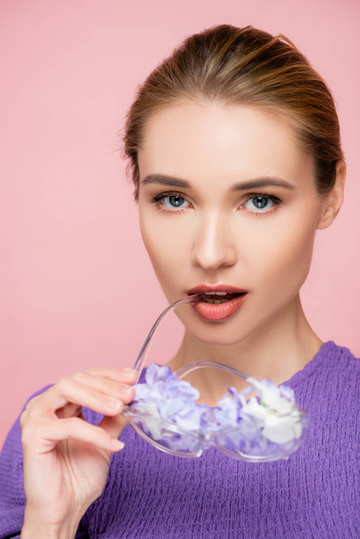 jonge vrouw met natuurlijke make-up houden bril met bloemen in de buurt van lippen geïsoleerd op roze - Foto, afbeelding