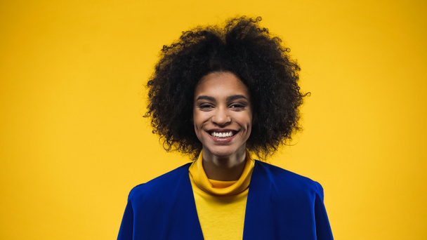 Mulher americana africana positiva em blazer azul olhando para a câmera isolada no amarelo - Foto, Imagem