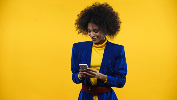 glimlachen Afrikaans amerikaanse vrouw in blauwe jas met behulp van smartphone geïsoleerd op geel - Foto, afbeelding