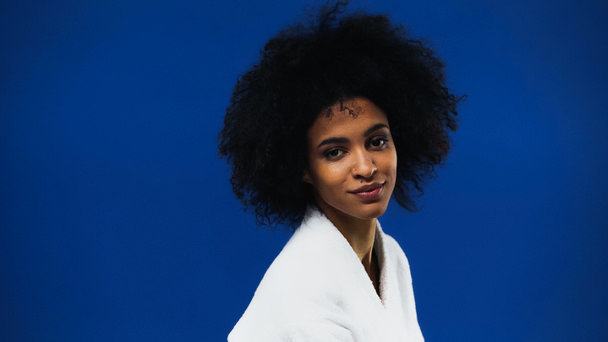 Young african american woman in white bathrobe looking at camera isolated on blue  - Фото, зображення
