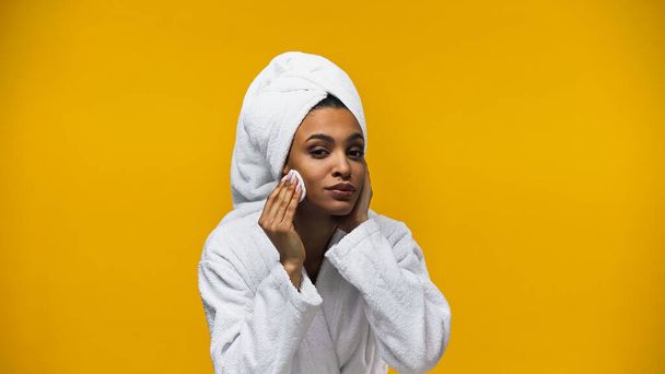 Αφρικανή Αμερικανίδα με πετσέτα και μπουρνούζι καθαρισμού προσώπου με βαμβακερό μαξιλάρι που απομονώνεται στο κίτρινο  - Φωτογραφία, εικόνα