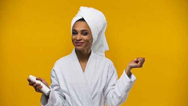 Χαρούμενη Αφροαμερικανή γυναίκα με πετσέτα και μπουρνούζι που κρατάει αποσμητικό και απομονώνεται στο κίτρινο.  - Φωτογραφία, εικόνα