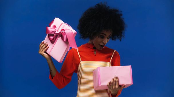 Mujer afroamericana asombrada abriendo regalo aislado en azul - Foto, imagen