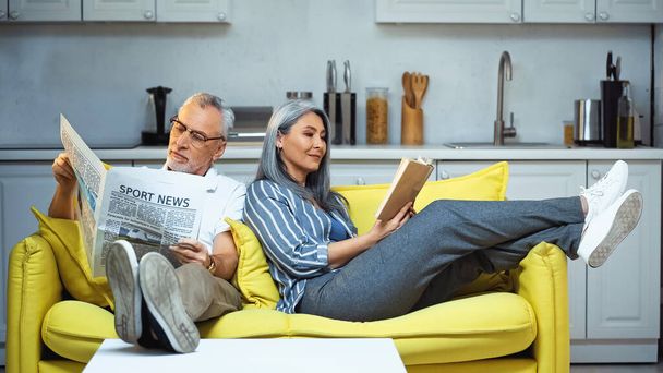 ηλικιωμένοι διαφυλετικό ζευγάρι ανάγνωση βιβλίο και εφημερίδα στον καναπέ στο σπίτι - Φωτογραφία, εικόνα