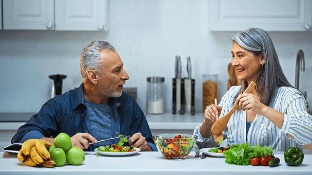 boldog idős fajok közötti pár beszél reggeli közben a konyhában - Fotó, kép