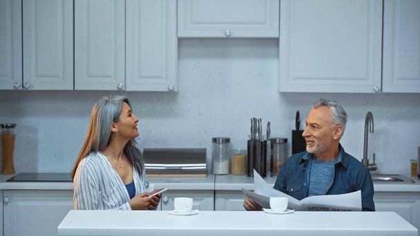 elderly interracial couple talking during morning coffee in kitchen - Foto, afbeelding