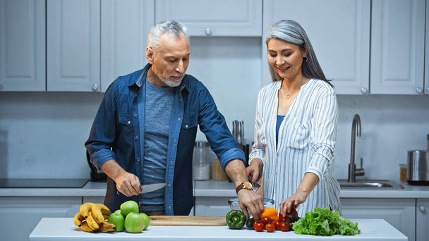 mosolygó fajok közötti pár készít reggelit friss zöldségekkel és gyümölcsökkel a konyhában - Fotó, kép