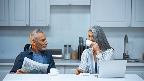 grey haired asian woman drinking coffee near husband with newspaper in kitchen - Foto, Imagem