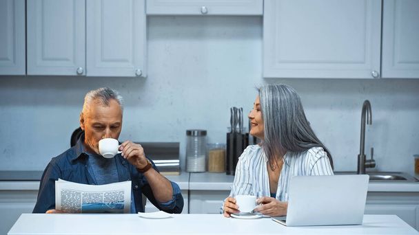 sonriente asiático mujer hablando a anciano marido bebiendo café y leyendo periódico en cocina - Foto, imagen