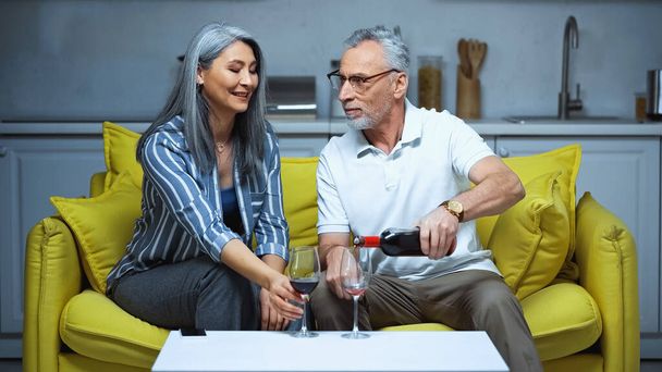senior uomo versando rosso vino vicino sorridente asiatico moglie su divano a casa - Foto, immagini