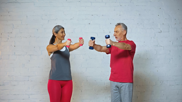 pareja interracial senior mirándose mientras hacen ejercicio con pesas  - Foto, Imagen